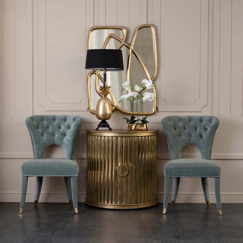Decorative side table with gold lamp and mirror, flanked by two blue chairs against a beige wall.