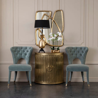 Decorative side table with gold lamp and mirror, flanked by two blue chairs against a beige wall.