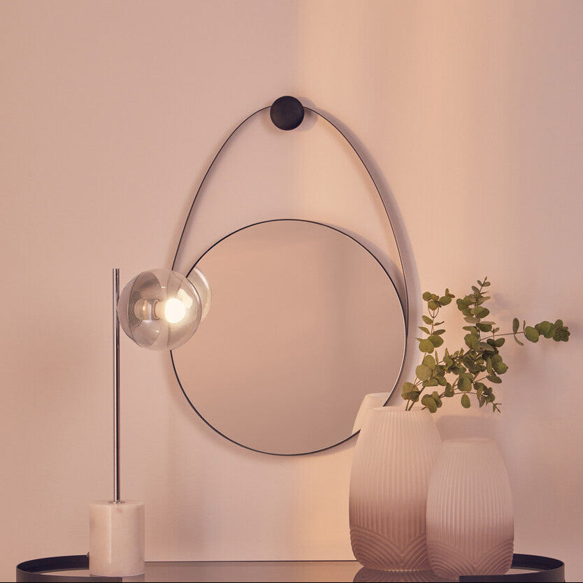 Modern interior with round mirror, lamp, and vases on a shelf against a light pink wall.
