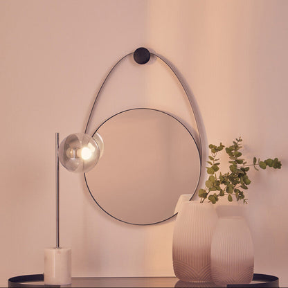 Modern interior with round mirror, lamp, and vases on a shelf against a light pink wall.