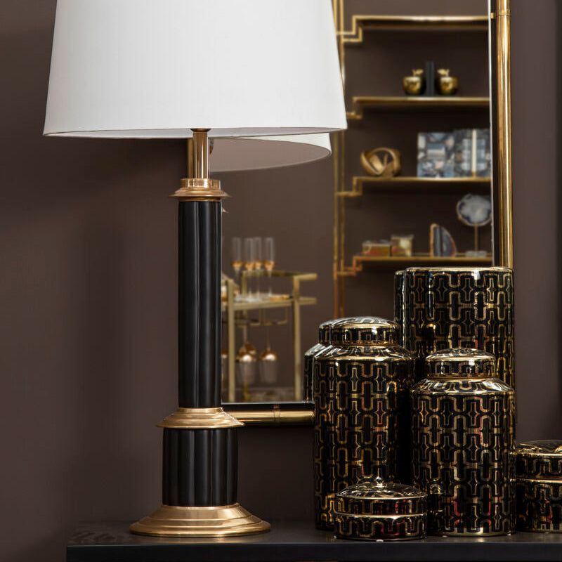 Decorative table lamp with a white shade and gold base in front of a mirror on a dark wall.