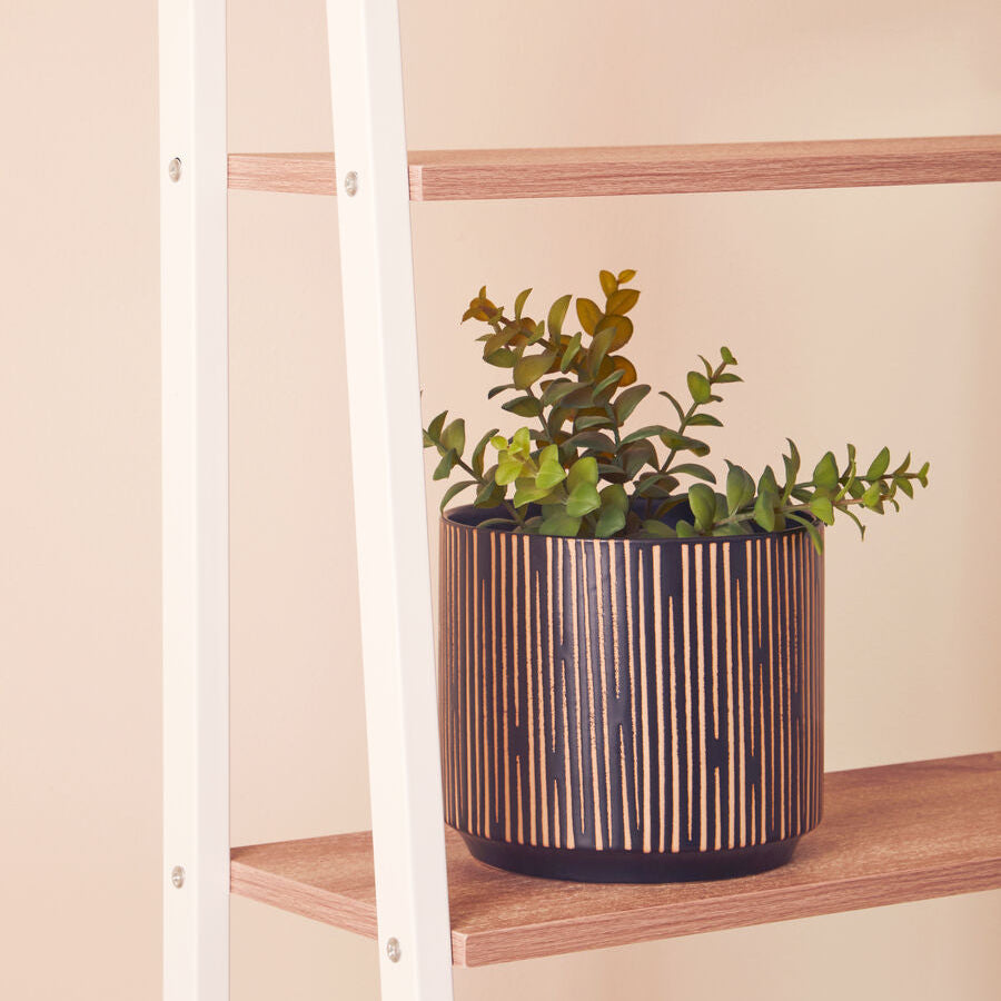 Plant in a decorative pot on a wooden shelf with a white ladder against a beige wall.