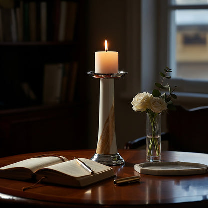 Candle on a wooden stand with an open book, pen, and vase of flowers on a table.