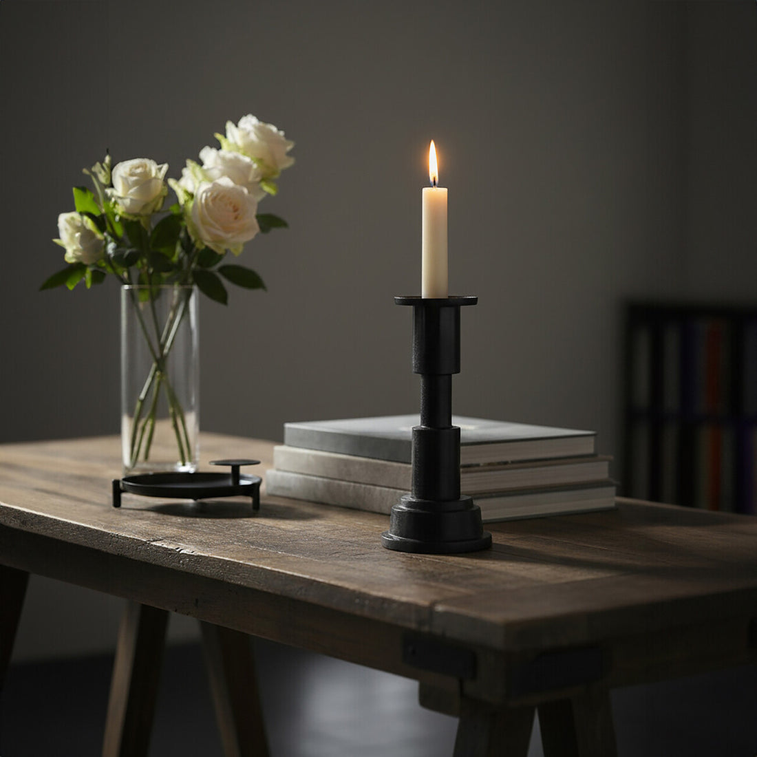 Candle in a black holder on a stack of books with white roses in a vase on a wooden table.