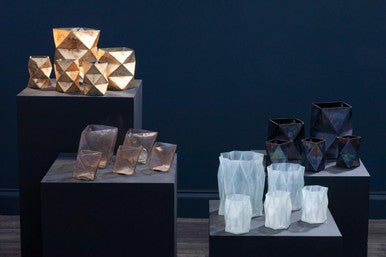 Collection of geometrically shaped vases on black pedestals against a dark blue wall.