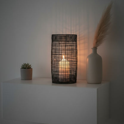Decorative lamp with a candle inside on a white surface, with a vase and plant in the background.