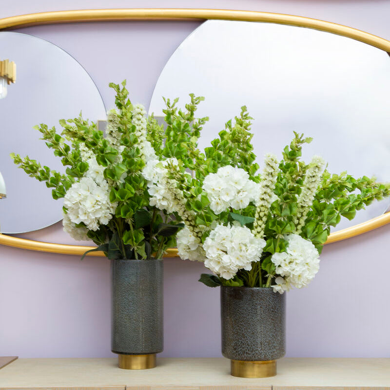 Two vases with white flowers on a beige cabinet with gold handles against a light purple wall.