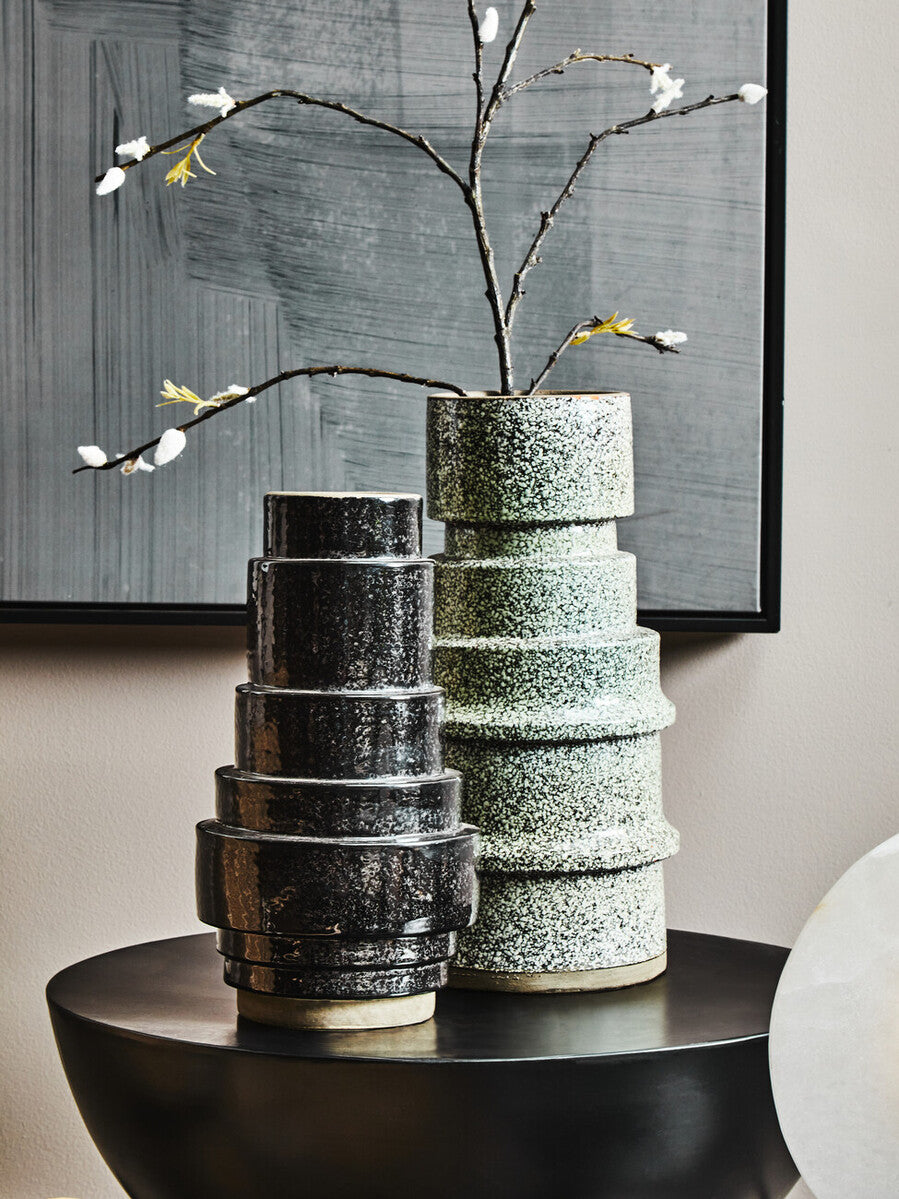 Stack of textured ceramic vases on a table with a branch and flowers against a neutral wall.