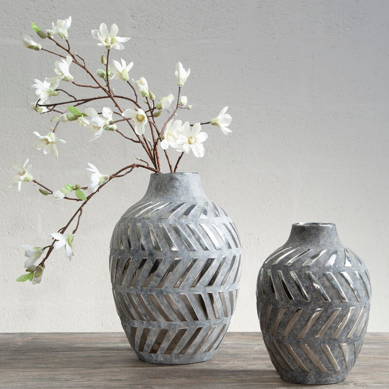 Two gray textured vases with a branch of white flowers on a wooden surface.