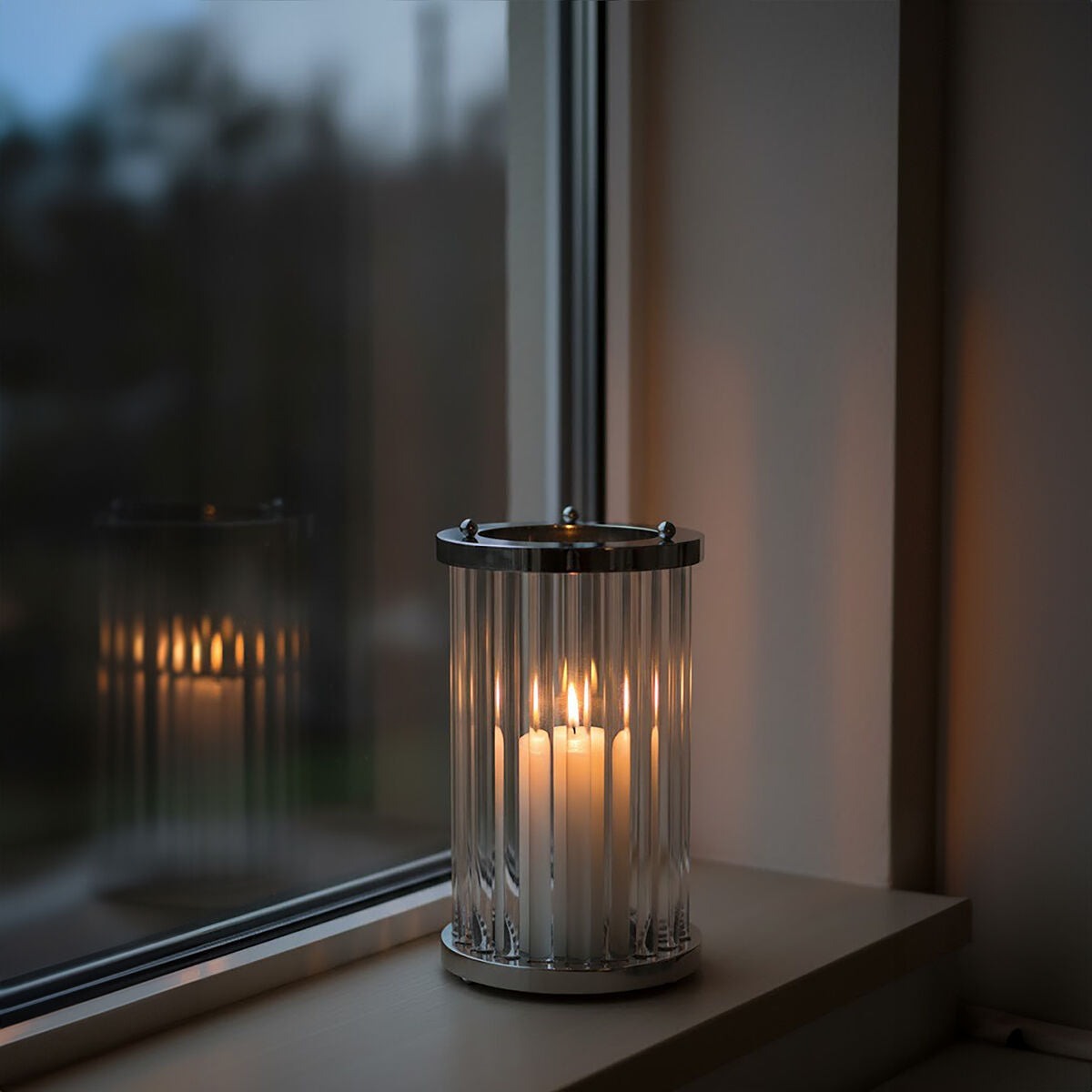 Glass candle holder with a lit candle on a windowsill, reflecting in the glass.