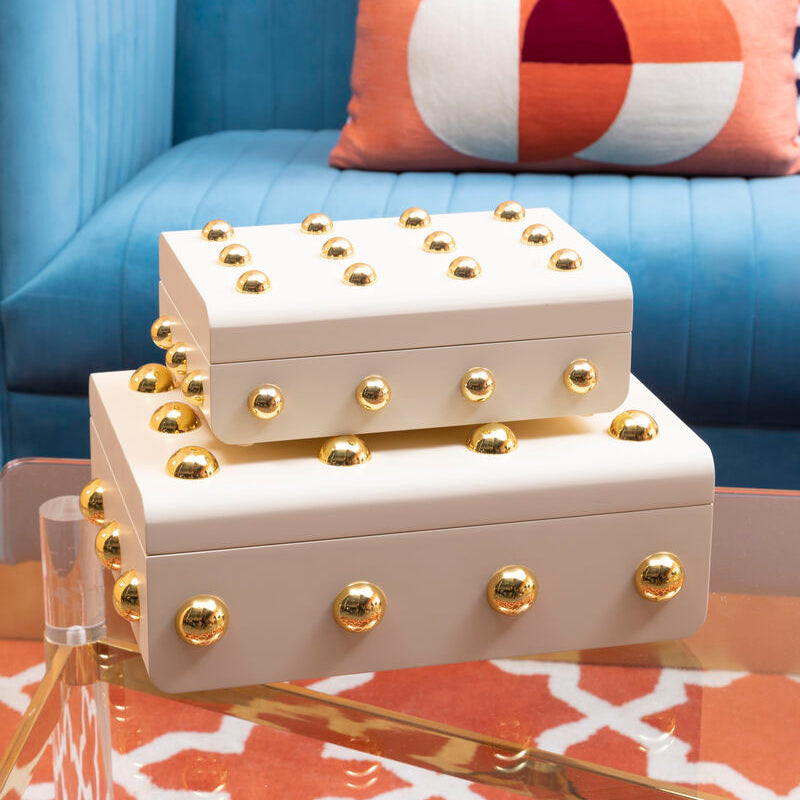 Two cream-colored boxes with gold embellishments on a coffee table in front of a blue couch with patterned cushions.