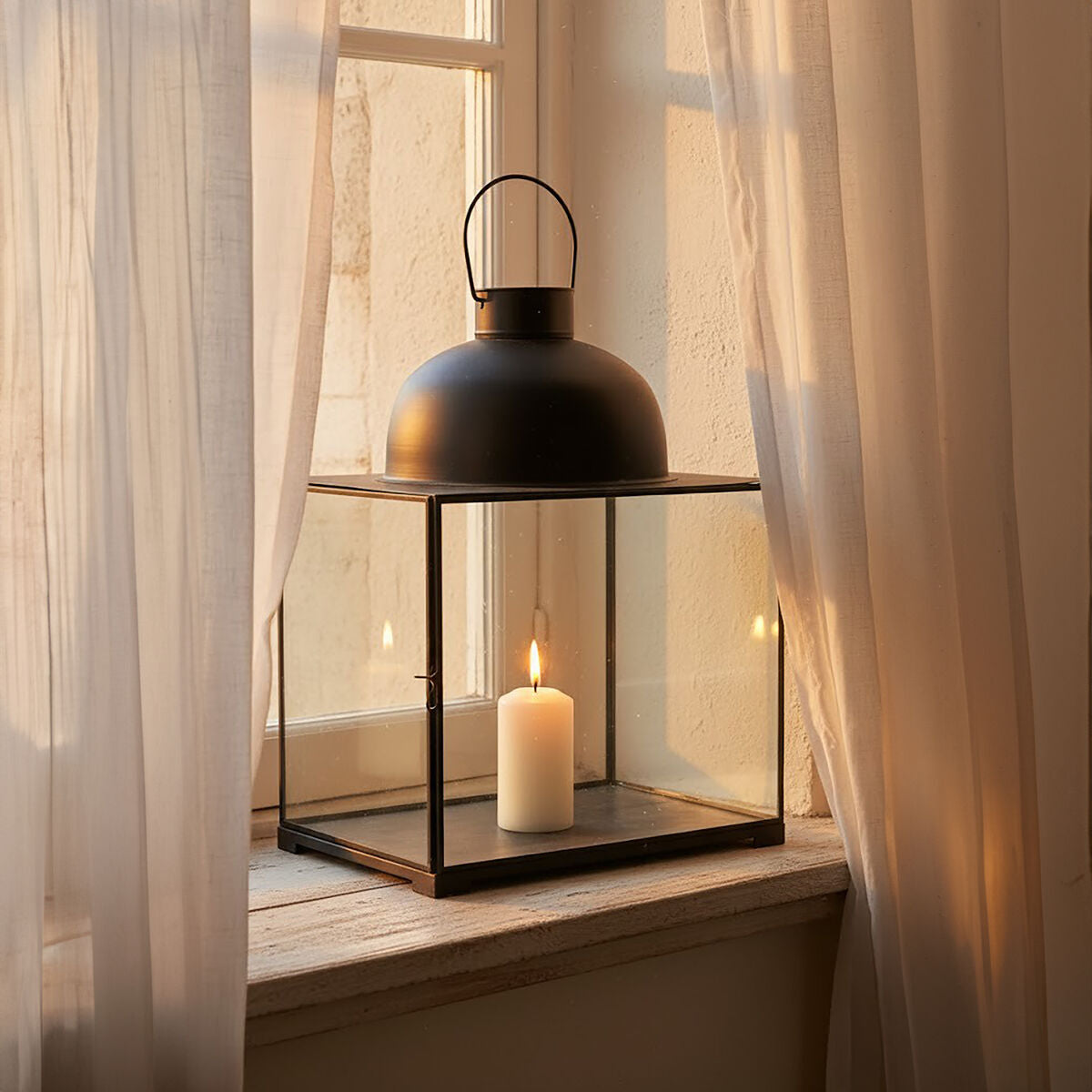 Black lantern with a lit candle on a windowsill with sheer curtains