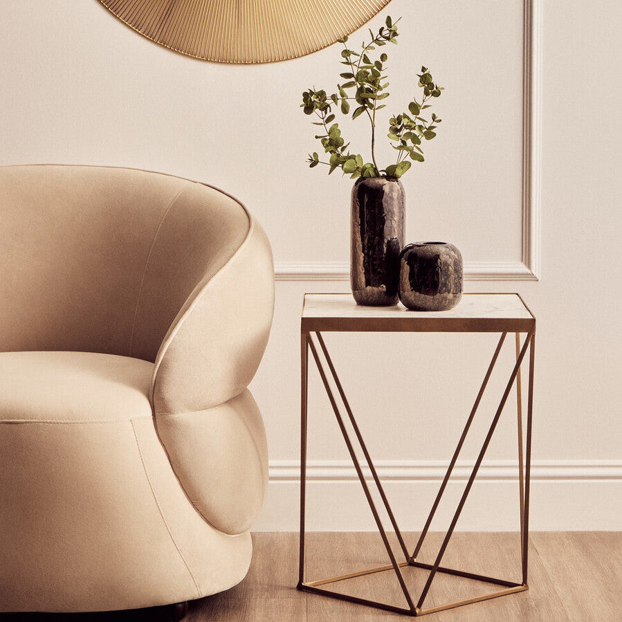 Beige armchair next to a small side table with decorative vases and a plant, against a neutral wall.
