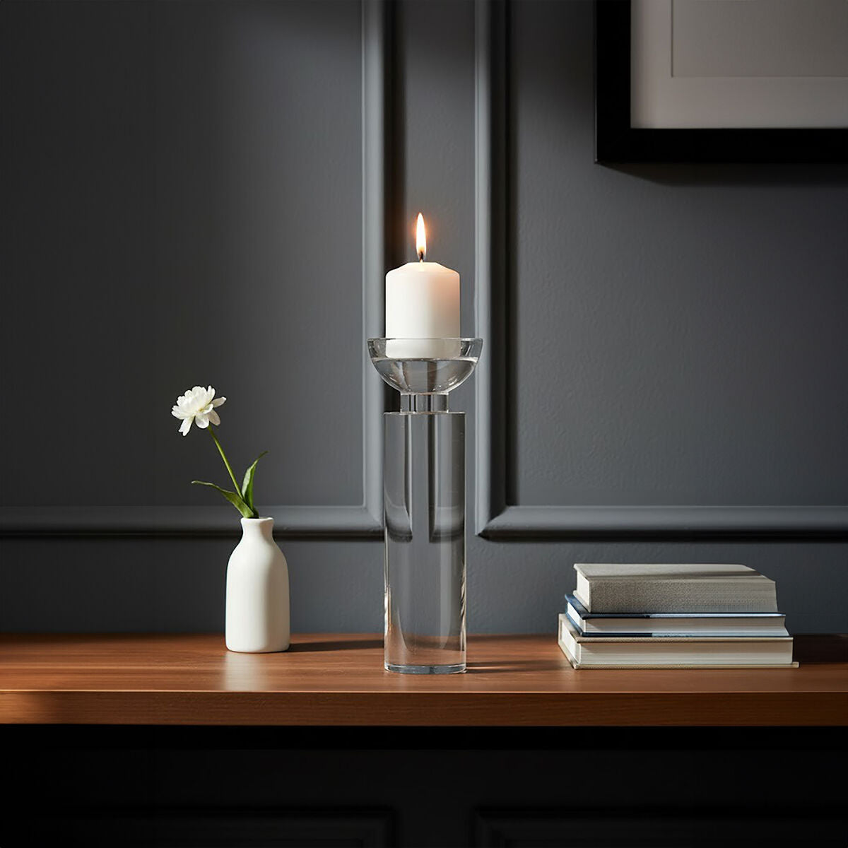 Candle in a clear glass holder with a white flower in a small vase on a wooden surface against a dark wall.