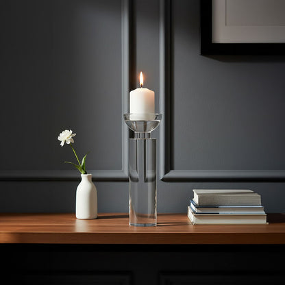 Candle in a clear glass holder with a white flower in a small vase on a wooden surface against a dark wall.
