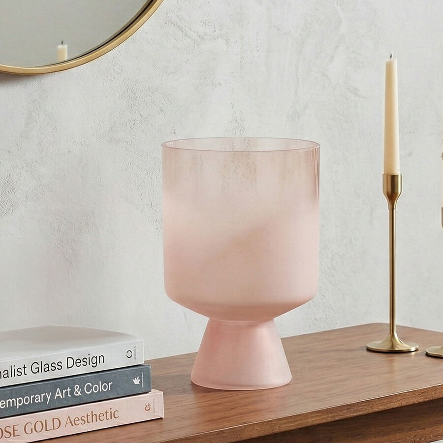 Pink glass vase on a wooden surface with books and a candle in the background.