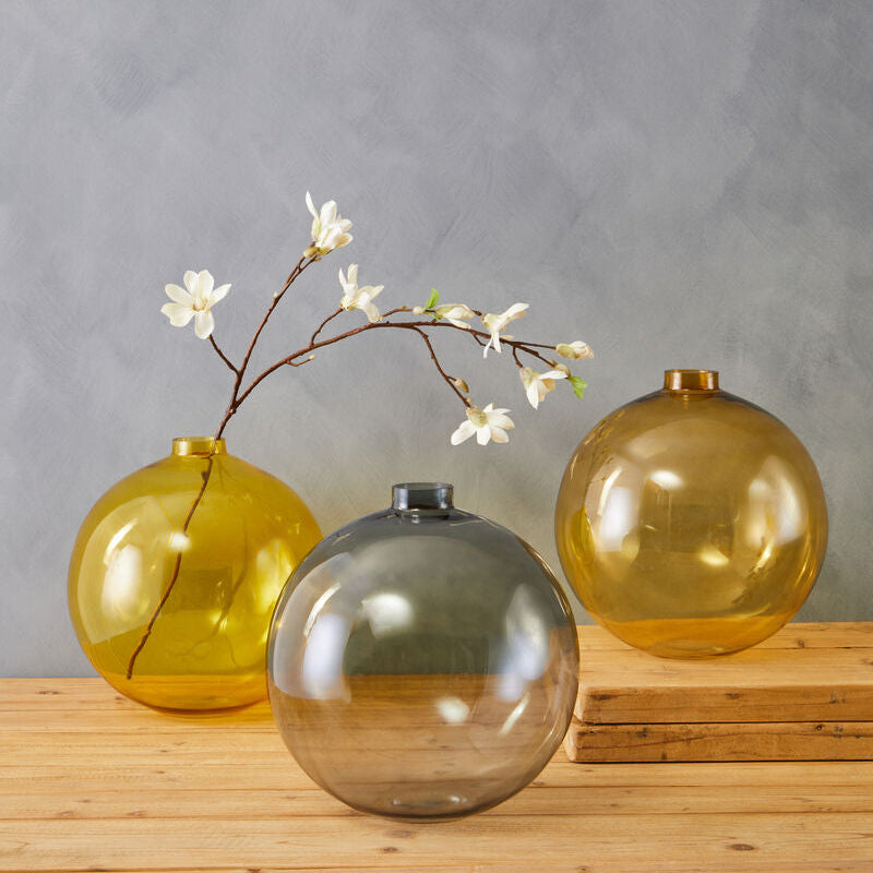 Three glass vases in yellow, green, and amber on a wooden surface with a gray background.