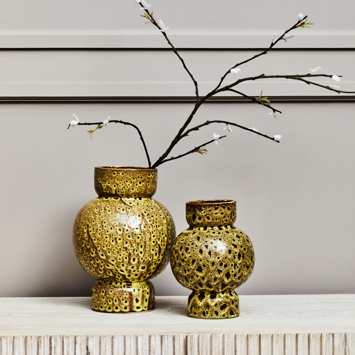 Two gold textured vases on a surface with decorative branches against a neutral background