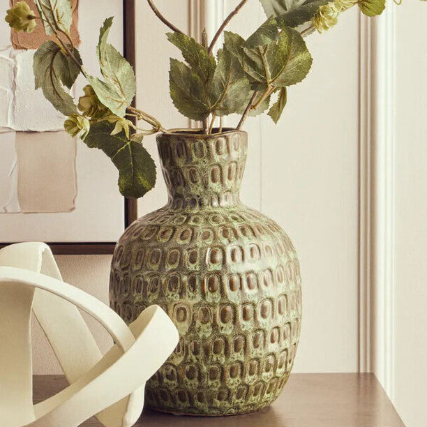 Decorative vase with green leaves on a wooden surface in a room with a colorful abstract painting.