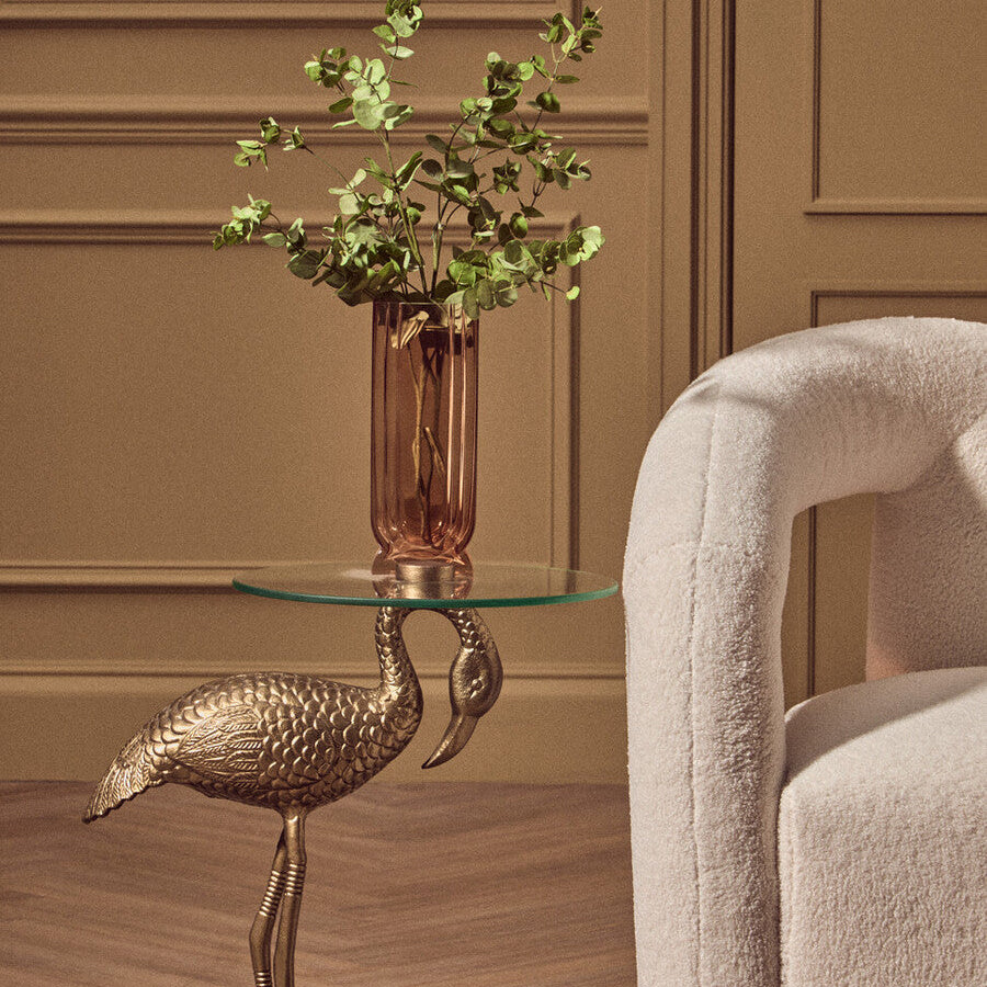Gold flamingo table with a glass top holding greenery, next to a beige armchair.