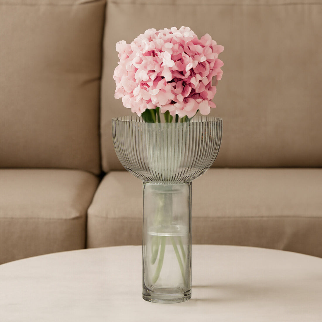 Clear glass vase with pink flowers on a beige surface with a beige sofa in the background