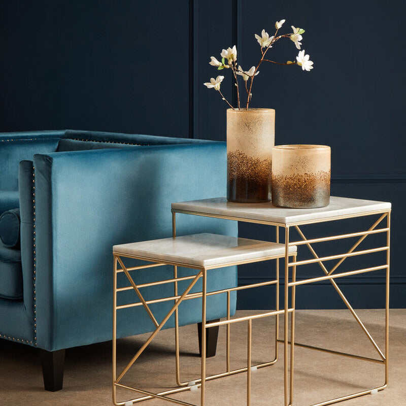 Gold side table with marble top next to a blue armchair against a dark wall.