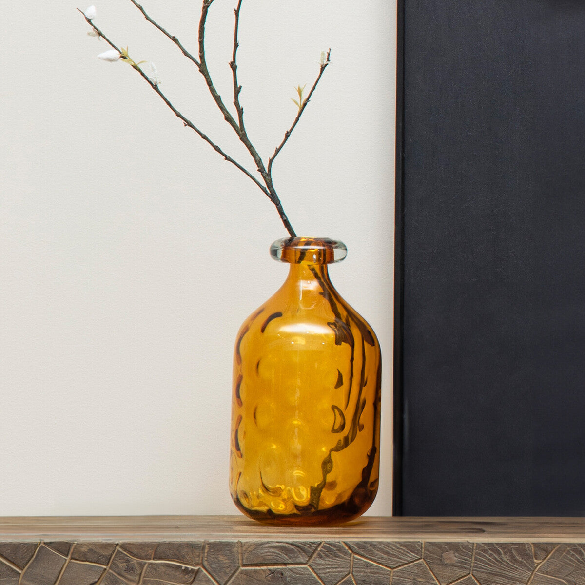 Amber glass vase with branches on a wooden shelf against a black and white wall.