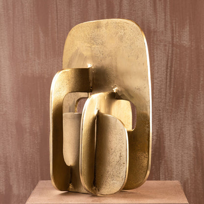 Gold abstract sculpture on a brown pedestal against a textured brown wall