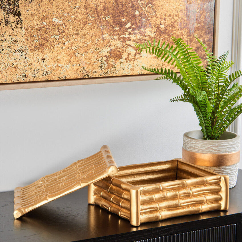 Decorative gold bamboo box on a black surface with a plant and abstract painting in the background.