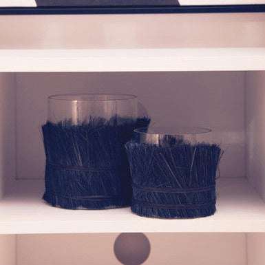 Two glass candle holders with navy blue fringe on a white shelf.