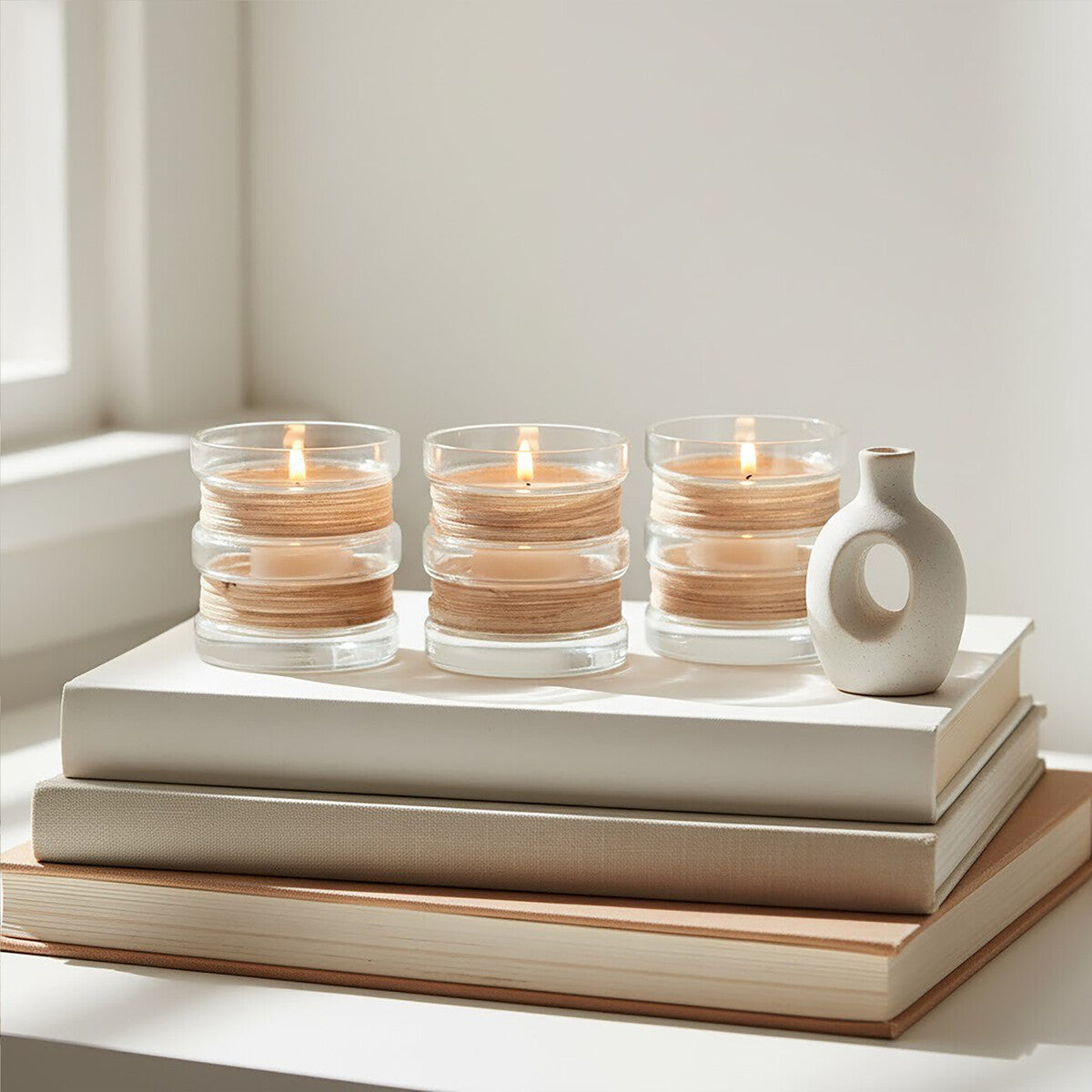 Three stacked candles in glass holders on books with a white vase beside them.