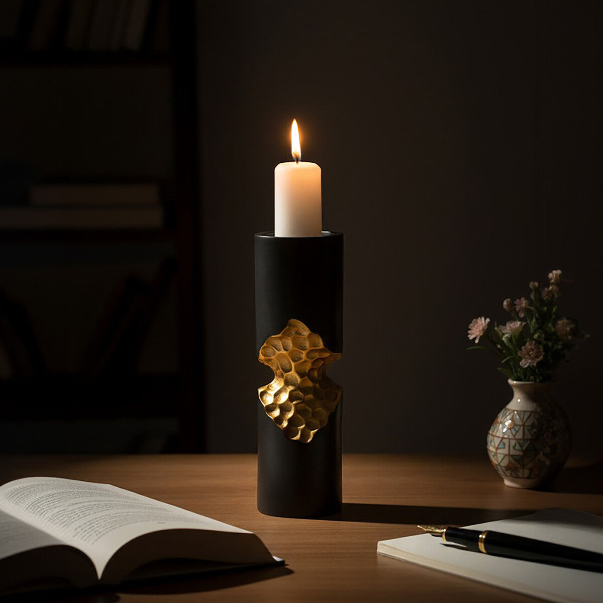 Candle in a decorative holder on a table with an open book and pen in a dimly lit room.