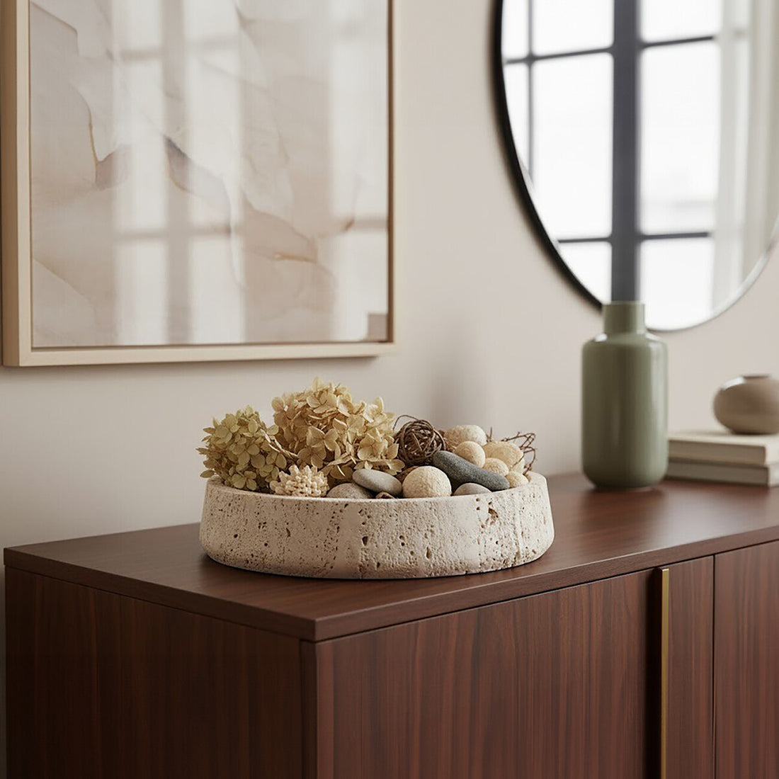 Decorative bowl with flowers and stones on a wooden surface, with a vase and framed artwork in the background.
