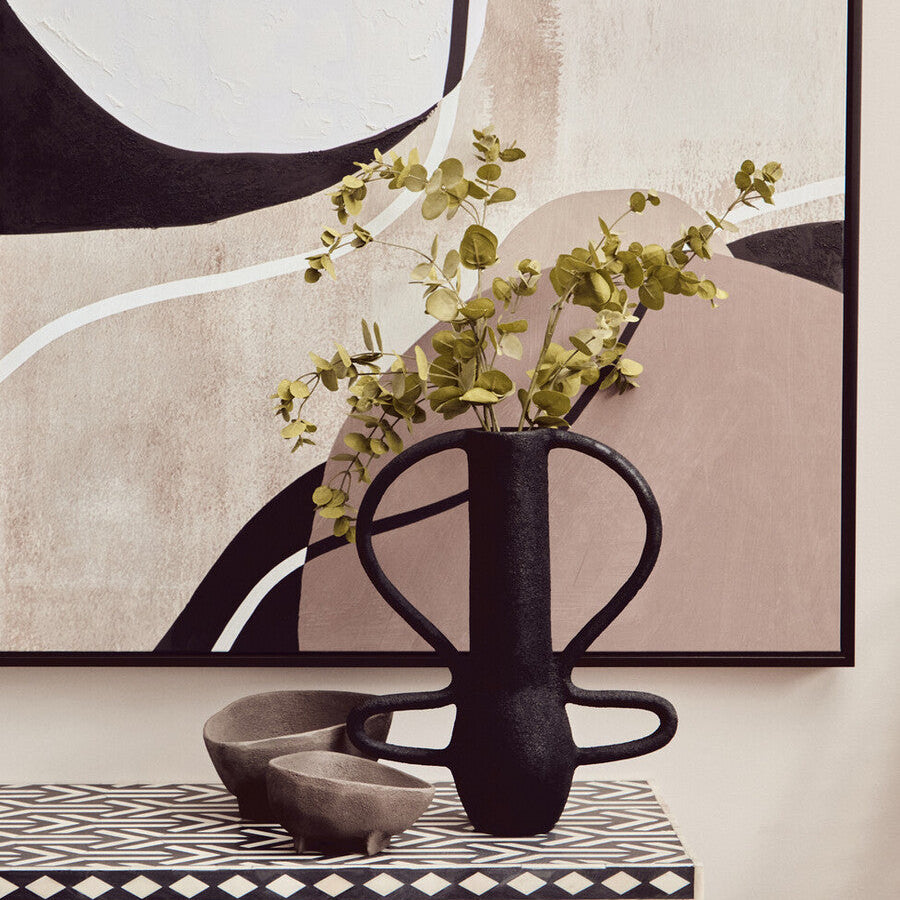 Black vase with greenery on a table in front of abstract wall art