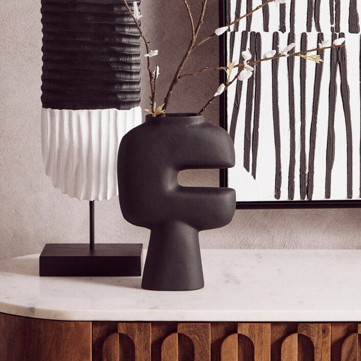 Decorative black vase with branches on a surface, with a lamp and abstract art in the background.