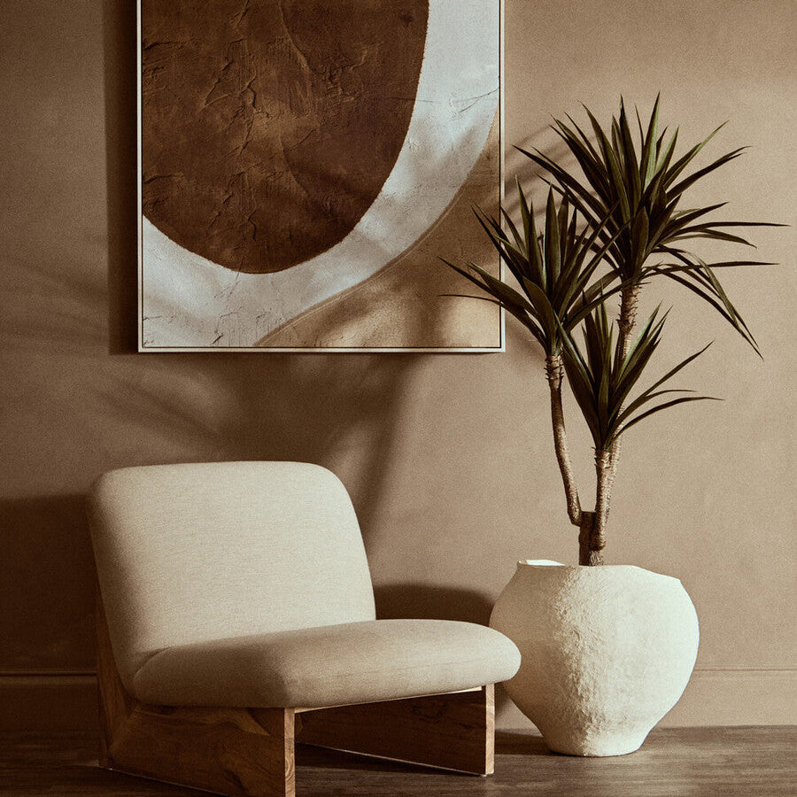 Beige armchair next to a potted plant with abstract art on the wall in a warm-toned room.
