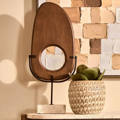 Decorative table with a wooden mirror, textured vase, and plant against a tiled wall.