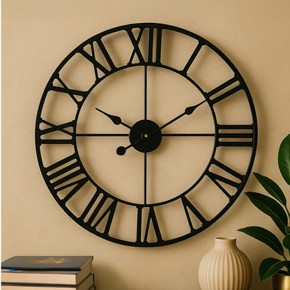 Large black wall clock with Roman numerals on a beige wall, next to a vase and books.