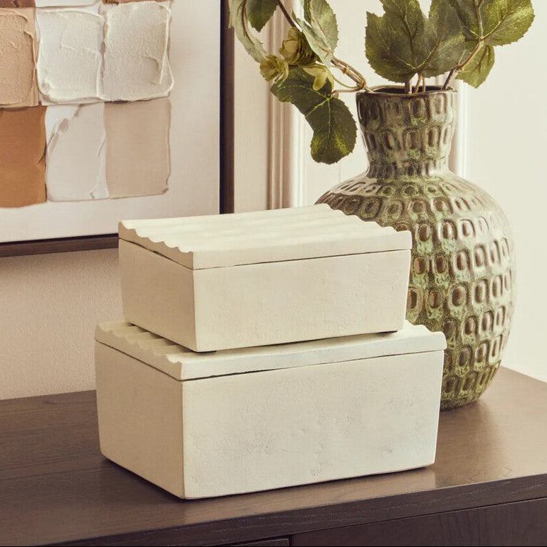 Two beige boxes stacked on a surface with a green textured vase and plant in the background.