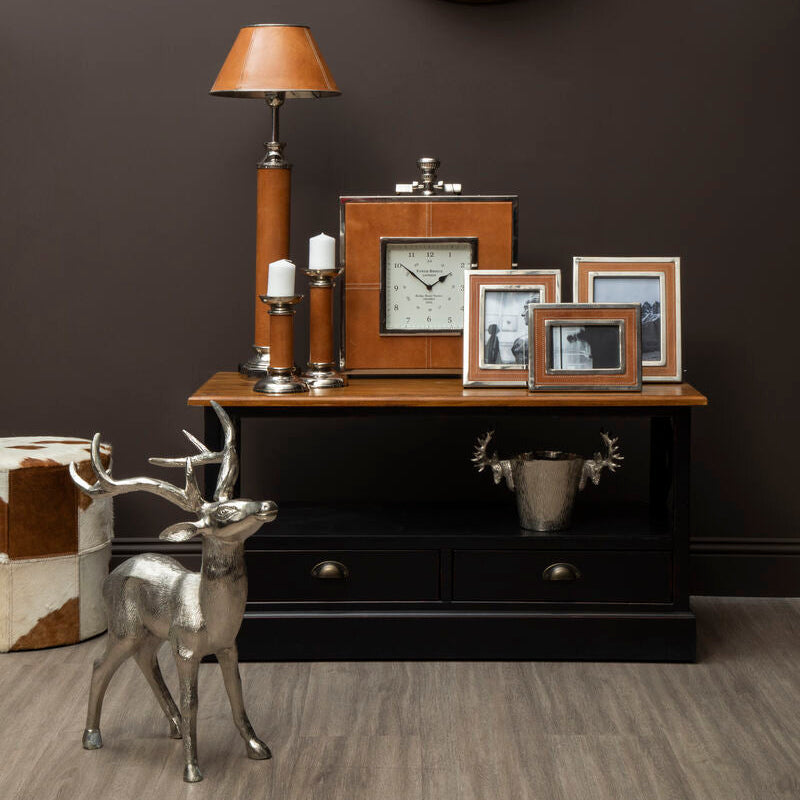 Decorative setting with a clock, lamp, and framed pictures on a wooden console against a dark wall.