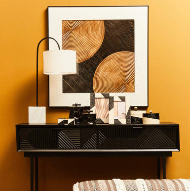 Modern interior design with a black console table, abstract art, and patterned stool against a yellow wall.