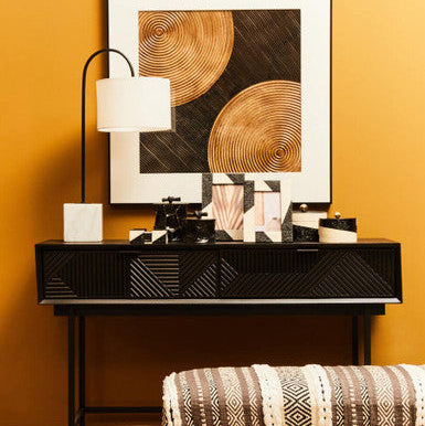 Modern interior design with a black console table, abstract art, and patterned stool against a yellow wall.