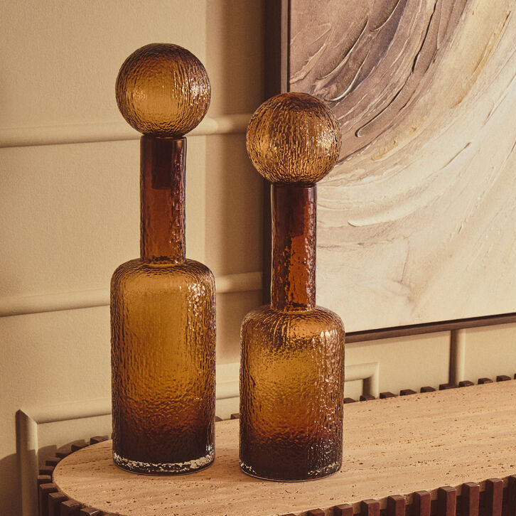 Two amber glass bottle-shaped decorative vases with spherical tops, displayed on a curved wooden console table in an interior setting.