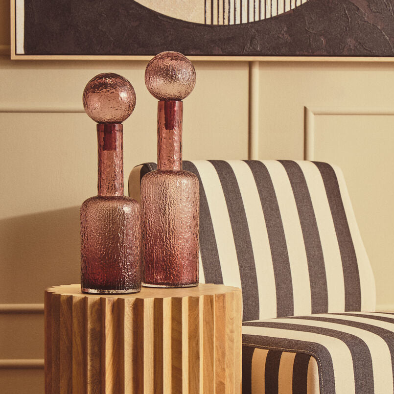 A modern cylindrical wooden side table with vertical fluting, holding two textured rose-gold glass bottle vases, set next to a black and cream striped armchair in a warm interior room.