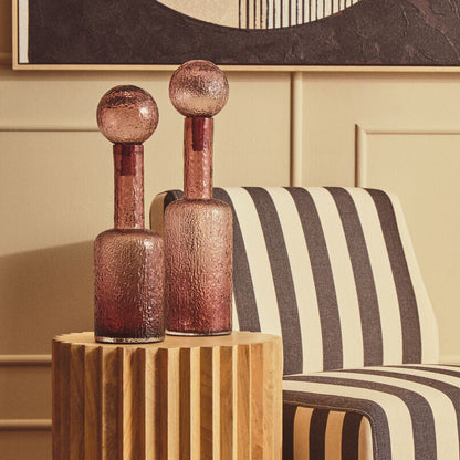 A modern cylindrical wooden side table with vertical fluting, holding two textured rose-gold glass bottle vases, set next to a black and cream striped armchair in a warm interior room.