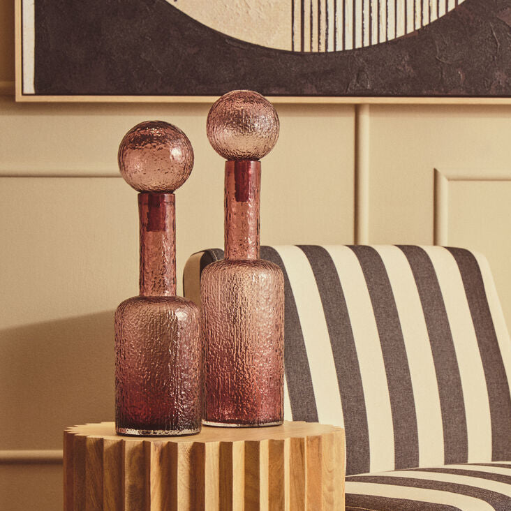 A modern cylindrical wooden side table with vertical fluting, holding two textured rose-gold glass bottle vases, set next to a black and cream striped armchair in a warm interior room.