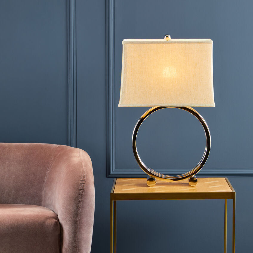 Table lamp with beige shade on a wooden side table against a blue wall with a pink armchair.