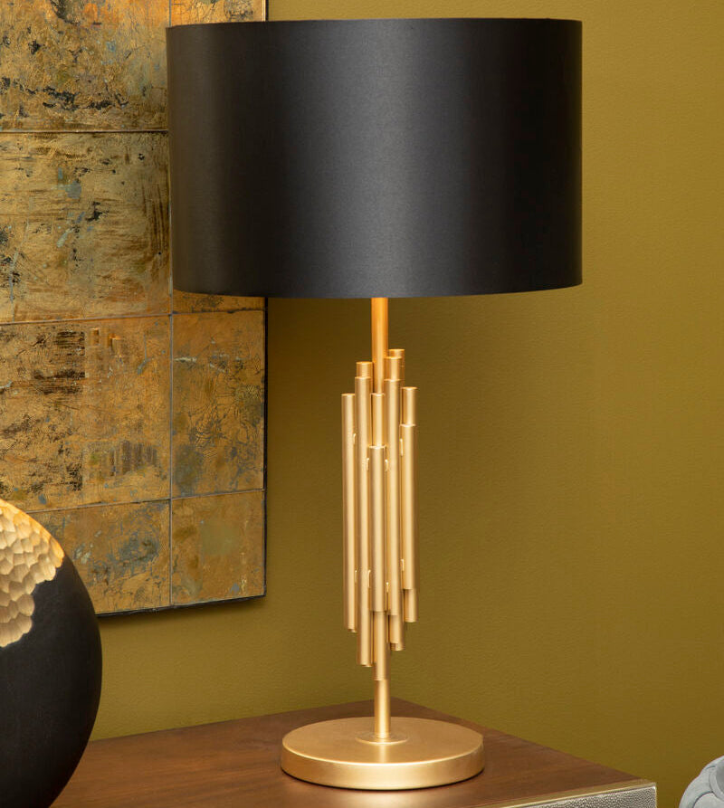 Table lamp with black shade and gold base on a wooden surface against a green wall.