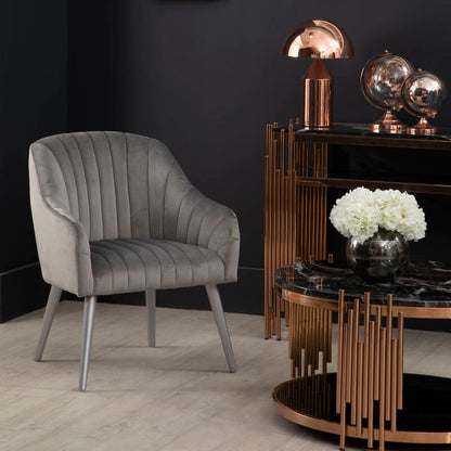 Gray velvet chair in a modern interior setting with a dark wall, decorative table, and lamp.