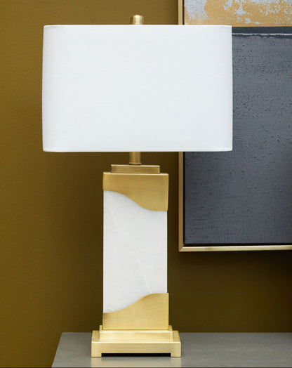 Table lamp with white shade and gold and marble base on a dark surface against a brown wall.